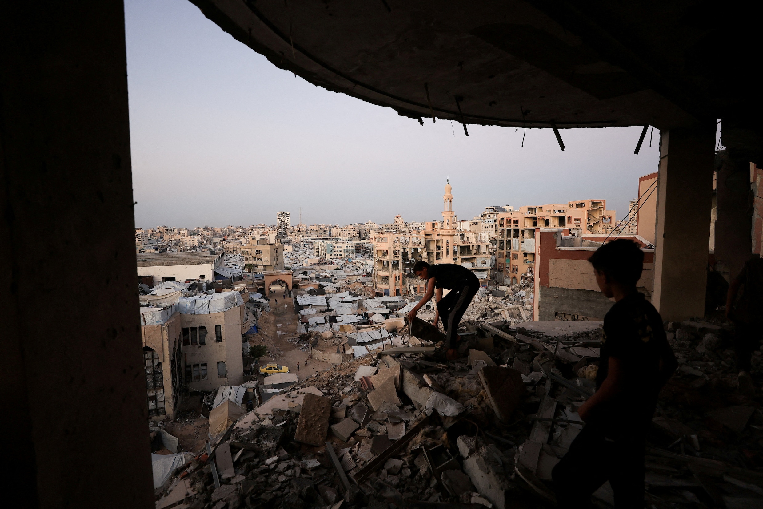 Een man zoekt in het puin van een gebombardeerd gebouw in Gaza Stad. Op de achtergrond is een tentenkamp voor ontheemden te zien. 